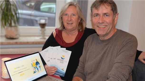 Eine Frau und ein Mann sitzen an einem Tisch. Die Frau hält Flyer von "Weser-Elbe hilft" in der Hand, auf dem Tisch steht ein Laptop, auf dem die Webseite der Organisation geöffnet sit.