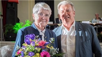 Ältere Frau mit Blumenstrauß steht lächelnd neben einem Mann in hellem Hemd und Sakko. Beide blicken in die Kamera, im Hintergrund läuft eine Veranstaltung.