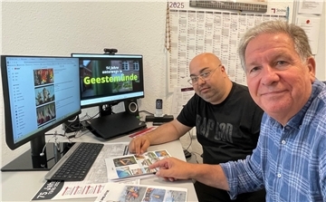 Zwei Männer, die an einem Tisch sitzen. Sie deuten auf ein buntes Blatt Papier. Im Hintergrund zeigt ein Computer-Bildschirm ein Video. Zu sehen ist der Satz "14 Jahre unterwegs in Geestemünde".