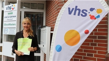 VHS-Leiterin steht vor dem Eingang eines Gebäudes und hält mehrere Programmhefte für das zweite Halbjahr 2025 in der Hand, neben einem VHS-Banner.
