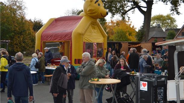 Leute stehen auf einem Platz an runden Bistrotischen und plaudern, im Hintergund eine rot-gelbe Hüpfburg.  