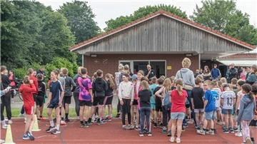 Viele Kinder, Jugendliche und Erwachsene in Sportkleidung stehen auf einer Laufbahn vor einem Gebäude