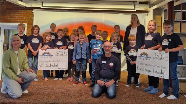 Eine Gruppe von Grundschulkindern aus Lunestedt steht gemeinsam mit zwei Erwachsenen vor einem bunten Hintergrund. Zwei große Spendenschecks über je 3200 Euro für das SOS-Kinderdorf und das Kinderhospiz Löwenherz werden präsentiert.