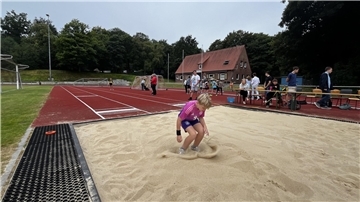 Ein Kind landet beim Weitsprung in der Sandgrube auf dem Sportplatz in Bad Bederkesa. Im Hintergrund beobachten Helfer und weitere Teilnehmende den Wettbewerb.