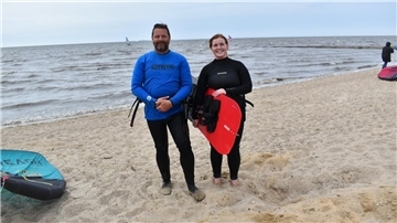 Ein Mann und eine Frau stehen am Strand. Die Frau trägt ein Surfbrett, im Hintergrund ist Wasser zu sehen.