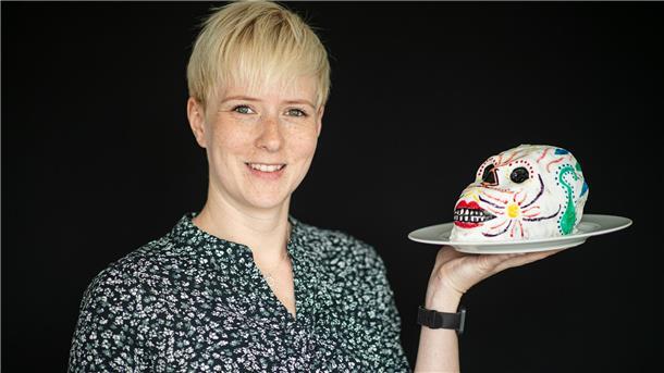 Eine Frau hält einen bunten Totenschädelkuchen auf einem Teller in der Hand und lächelt in die Kamera.