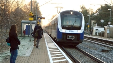 Ein Zug an einer kleineren Haltestelle. Auf dem Bahnsteig warten Menschen auf den Zug.  