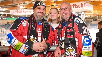 Fans lächeln in die Kamera am Rande eines Eishockeyspiels.
