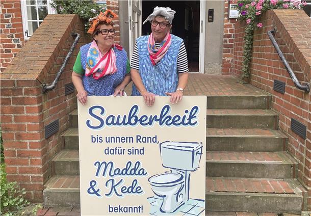 Zwei als Hausfrauen verkleidete Frauen stehen auf einer Treppe vor der Burg zu Hagen und halten ein großes Schild mit der Aufschrift: „Sauberkeit bis unnern Rand, dafür sind Madda & Kede bekannt!“