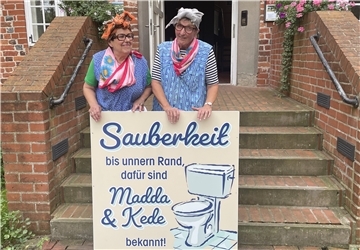 Zwei als Hausfrauen verkleidete Frauen stehen auf einer Treppe vor der Burg zu Hagen und halten ein großes Schild mit der Aufschrift: „Sauberkeit bis unnern Rand, dafür sind Madda & Kede bekannt!“