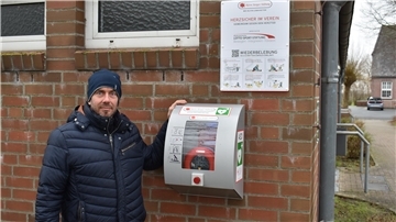 Vorstandsmitglied Ralf Buhrmann zeigt den jetzt betriebsbereiten Defibrillator an der Westseite der Sporthalle in Esenshamm.
