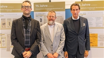 Drei Männer stehen nebeneinander, dahinter Schautafeln mit Grafiken zur Entwicklung der philosophischen Gesellschaft Bremerhaven