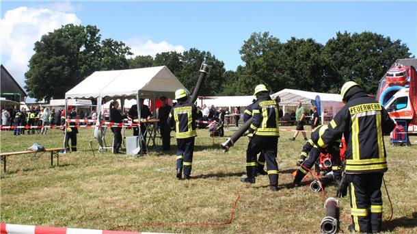 Feuerwehrleute in Einsatzkleidung führen bei einem Leistungsvergleich auf einer Wiese Übungsaufgaben mit Schläuchen und Geräten durch, während im Hintergrund Zuschauer unter Pavillons stehen.