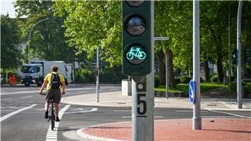 Eine Straßenkreuzung: Im Vordergrund steht eine Fahrradampel mit Grünlicht. Im Hintergrund eine Fahrradampel mit Rotlicht. Auf dem Fahrradweg ist ein Radfahrer unterwegs.