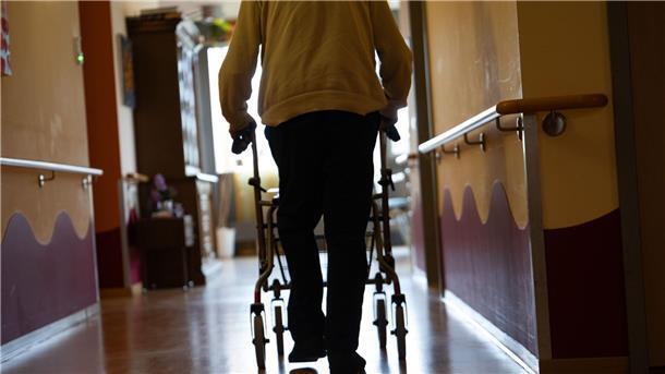 Eine ältere Person geht auf einen Rollator gestützt durch einen Flur eines Krankenhauses.