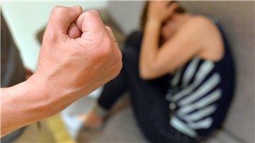 Ein Mann zeigt seine Faust, eine Frau sitzt verängstigt auf dem FJußboden und drückt sich an eine Wand.