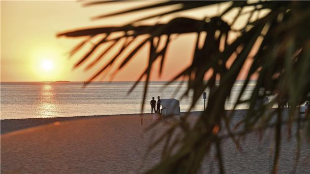 Zwei Menschen stehen neben einem Strandkorb am Strand und beobachten den Sonnenuntergang.