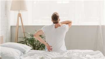 Ein Mann, der offensichtlich Rückenschmerzen hat, sitzt auf einem Bett.