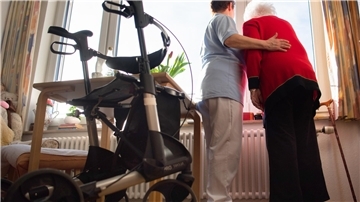 Zwei Frauen stehen an einem Fenster und im Vordergrund steht ein Rollator.