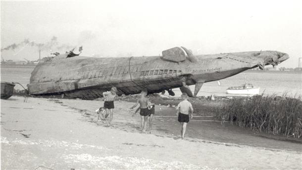 Wie ein abgestürztes Raumschiff liegt es am Strand von Blexen: Das Heck eines deutschen U-Bootes - 1945 von der Besatzung selbst in der Außenweser versenkt, einige Jahre später von mutigen Männern des Schrottwerts wegen wieder geborgen. Kinder spielen davor - ein Bild, in dem sich die 50er Jahre des Nachkriegsdeutschlands verdichten. Bei dem Boot dürfte es sich um U 38, ein Boot vom Typ IX A, gehandelt haben - 1938 in Dienst gestellt und 1.032 Tonnen schwer. Das Foto entstand im August 1955.