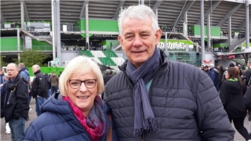 Ein älteres Ehepaar in blauen Daunenjacken steht vor einem Fußballstadion.