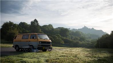 Ein VW Bulli steht auf einer Wiese vor einem Wald. Im Hintergrund ist eine Kirche auf einem Berg zu sehen.