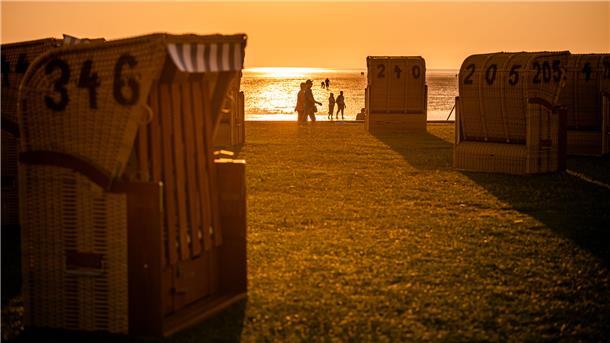 Im Vordergrund stehen Strandkörbe auf einem Grünstrand, im Licht des Sonnenuntergangs sind Silhouetten von Menschen zu sehen.