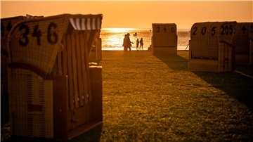 Im Vordergrund stehen Strandkörbe auf einem Grünstrand, im Licht des Sonnenuntergangs sind Silhouetten von Menschen zu sehen.
