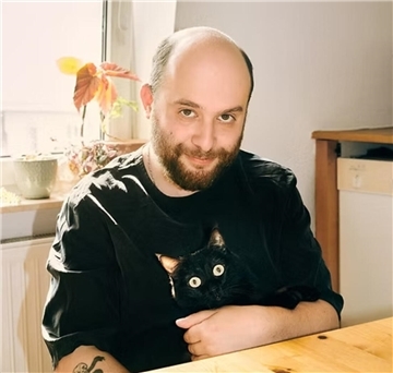Ein Mann sitzt an einem Tisch, auf seinem Schoß sitzt eine schwarze Katze.
