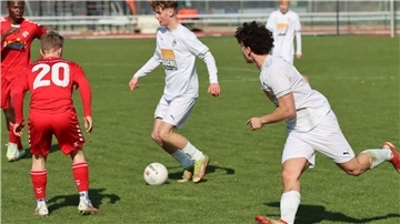 Zwei junge Männer in weißen Trikots laufen hinter einem Ball her, zwei junge Männer in roten Trikots stellen sich in den Weg.