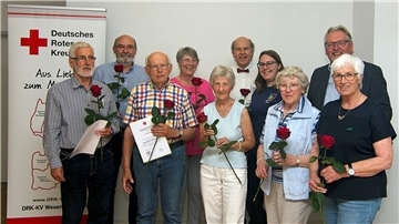 Gruppe von zehn Personen, die beim DRK geehrt wurden, stehen mit roten Rosen und Urkunden in der Hand vor einem Banner des Deutschen Roten Kreuzes.