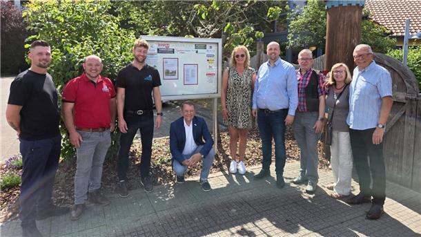 Eine Gruppe von Sponsoren und Organisatoren steht bei sonnigem Wetter vor dem neuen Informationsschaukasten des EWW Kindergartens Nimmerland.