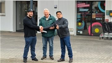 Drei Männer stehen vor einem Ladeneingang, zwei geben sich die Hand, einer zeigt Daumen hoch; alle lächeln, im Hintergrund Einkaufswagen und ein Schild mit Öffnungszeiten.