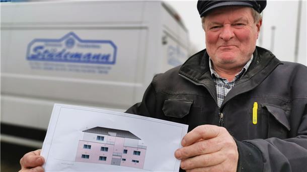 Ein Zimmermeister hält eine Grafik in den Händen, die einen Neubau zeigt. Im Hintergrund ist ein Kastenwagen zu sehen.