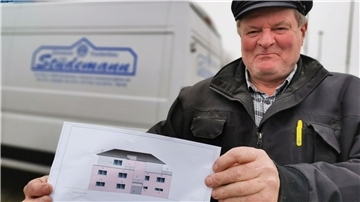 Ein Zimmermeister hält eine Grafik in den Händen, die einen Neubau zeigt. Im Hintergrund ist ein Kastenwagen zu sehen.