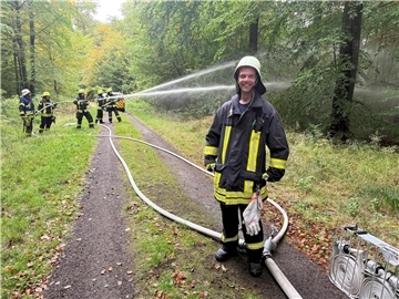Lachender Feuerwehrmann im Wald, dahinter Feuerwehrleute mit Schläuchen