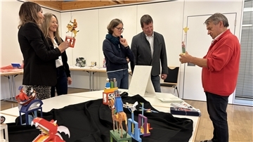 Papierkünstler Walter Ruffler zeigt mehreren interessierten Erwachsenen bunte mechanische Papierfiguren auf einer Tischpräsentation in einem Ausstellungsraum.