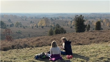 Zwei Personen sitzen auf einer Wiese inmitten einer offenen Heidelandschaft. Vor ihnen liegt eine weite Landschaft mit Bäumen, Sträuchern und sanften Hügeln, die in herbstlichen Farben leuchtet. Der Himmel ist leicht bewölkt.