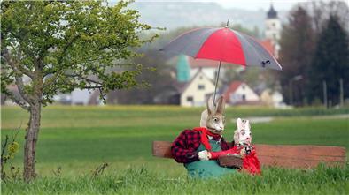 Zwei als Osterhasen dekorierte Strohpuppen sitzen mit einem großen Regenschirm an einem Feld.