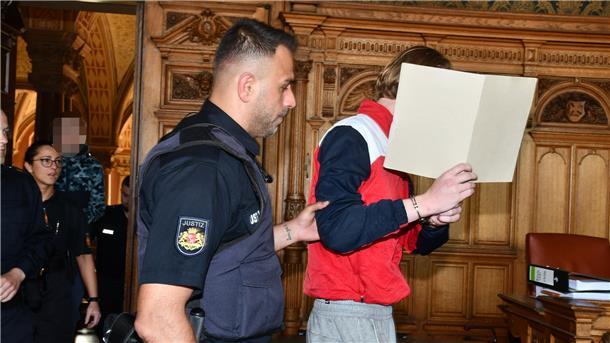 Chat unter Jugendlichen endet fast tödlich mit Messerattacke in Bremerhaven Angeklagter wird in Gerichtssaal geführt und hält eine Mappe vor das Gesicht.