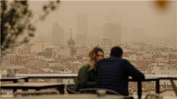 Zwei Menschen sitzen in Madrid auf einer Terrasse, während eine Wolke mit Sahara-Staub über die spanische Hauptstadt zieht. Die typische Auswirkung von Sahara-Staub ist ein roter oder orangener Himmel (Archivbild).