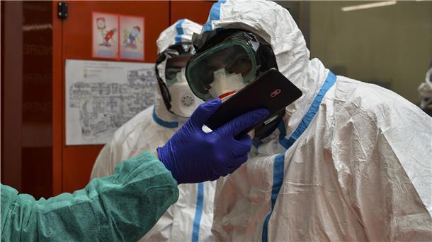 Zwei Mitarbeiter in Schutzanzügen schauen in einem Krankenhaus auf das Smartphone eines weiteren Angestellten.