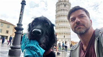 Ein Hund mit einem Tuch im Mund und ein Mann mit Bart schauen in die Kamera. Im Hintergrund steht der Schiefe Turm von Pisa.