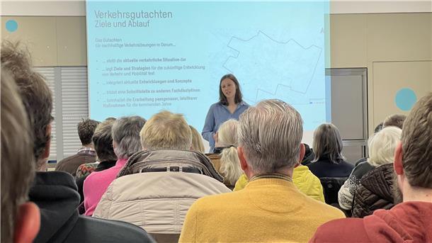 Im Hintergrund steht eine Frau mit langen dunkelblonden Haaren und im blauen Hemd. Sie hält einen Vortrag. Im Hintergrund ist eine Präsentation zu sehen. Im Vordergrund sitzen Menschen.